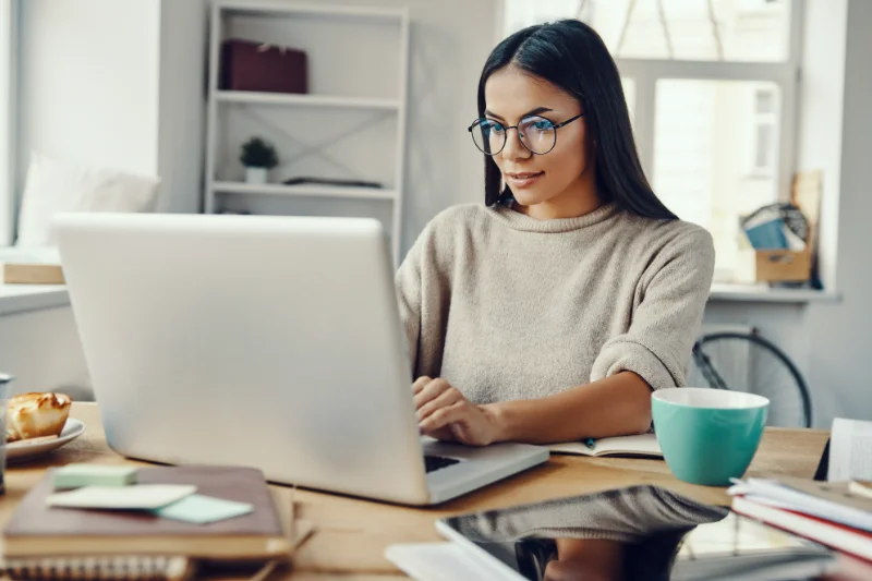 A woman with long dark hair and glasses is sitting at a desk, working on a laptop. She is surrounded by notebooks, a mug, and office supplies in a bright, modern workspace.