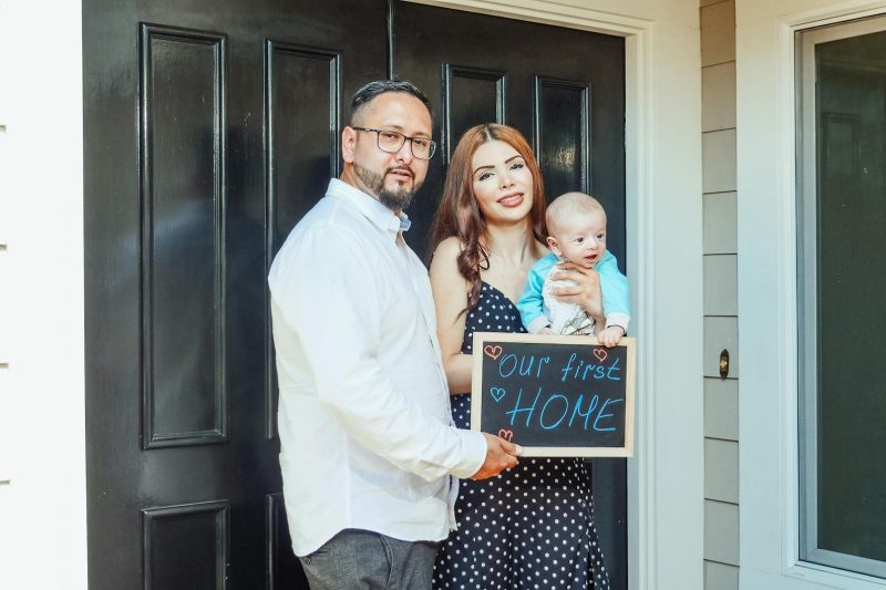 Home 136 Young family with baby holding a sign Our First Home